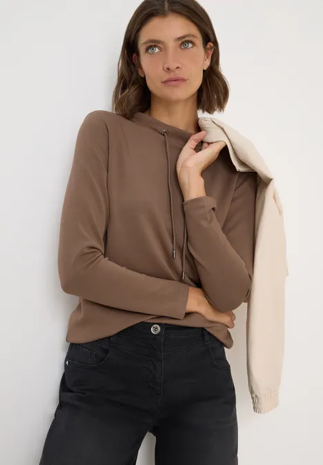 Frau mit braunem Hoodie und dunkler Jeans hält eine helle Jacke über der Schulter.